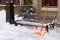 An orange snow shovel stands near a wooden bench