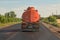 Orange tank truck rides on a country road against a blue sky. Back view
