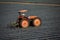 Orange tractor driven by man in straw hat pulling cultivator in strawberry field covered with plastic mulch