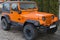 Orange two door jeep Wrangler SUV with gray roof and square headlights standing on gray sidewalk tiles in front of house