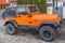 Orange two door jeep Wrangler SUV with gray roof and square headlights standing on gray sidewalk tiles in front of house