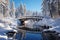 An ornate wooden bridge spans a calm, icy river, surrounded by tall snow-laden pine trees under a clear blue sky.