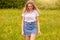 Ourdoor shot of young beautiful Caucasian girl in white t shirt and blue denim skirt posing against sunset in meadow, looking down