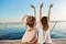 Outdoor back shot of two young female on luxury vacation, waving at seaside while sitting on yacht. Best friends are