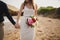 Outdoor beach wedding ceremony near the ocean, close up of hands of stylish couple with wedding bouquet, bride is