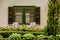 Outdoor conversation sitting area patio furniture in front of shuttered window and behind landscaped garden plants including hedge