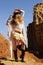 Outdoor fashion photo of young happy woman in hat, standigg against rocks, hands up