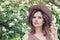 Outdoor lifestyle photo of young beautiful lady. Brown hair, fedora hat. Perfect face on blossoms flowers background