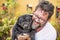 Outdoor nice portrait of adult caucasian man with black beard and same color old funny dog pug - garden in background and best