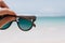 Outdoor photo of ocean with female hand on foreground. Woman holding summer sunglasses on sea background..