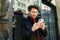 Outdoor photo of young brown haired unshaved guy keeping mobile phone in raised hands and looking positively on screen while