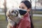 Outdoor picture of delighted lovely young female wearing antibacterial mask to protect from coronavirus, holding white Maltese dog