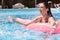 Outdoor picture of relaxed playful female having fun in swimming pool alone, lying on pink water mattress, splashing water, being