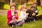 Outdoor portrait of positive inspired child, girl sitting on grass with legs crossed and using mobile phone in company