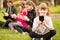 Outdoor portrait of positive inspired child, girl sitting on grass with legs crossed and using mobile phone in company