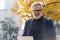 Outdoor portrait of sleek caucasian man with gray beard and black glasses, wearing checked blazer and black turtleneck