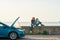 Outdoor shot of couple of travelers sitting near the broken car with open hood and smoking engine, man and woman looking