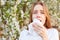 Outdoor shot of discontent young girl has seasonal allergy, uses tissue, poses over blooming tree, has rhinitis and sneezing,