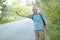 Outdoor shot of grey haired senior man in casual t shirt, has vacation trip, hitchhikes on road in countryside, has rucksack on ba