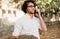 Outdoor shot of a smart young man talking on mobile phone in the street. Handsome male with curly hair in a white shirt and eyegla