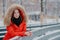 Outdoor shot of thoughtful European woman wears red winter jacket with hoody on head, leans on hence, looks pensively into