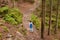 Outdoor shot of wandering female with ponytail wearing blue backpack, rose jacket and jeans, having walk in forest, choosing path