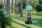 Outdoor shot of young brunette female practices yoga in green park holds fitness ball over head wears cropped top and leggings