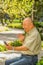 Outdoor view of old man sits on bench and saying hello on video cam with cellphone in his hand, enjoying the nature and