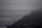 Outline of a fisherman on a foggy day sitting on a rocky outcropping holding a pole fishing at the beach