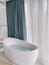 Oval bathtub with chrome taps stands near a panoramic window with curtains