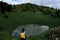 Oval mountain lake among alpine meadows. Girl traveler with dreadlocks in blue jacket with yellow backpack is enjoying life in