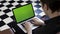 Over the shoulder view of a man using laptop with green screen in the room with chessboard floor. Stock footage. Male