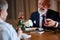 Over shoulder view of mature man in tuxedo proposing to woman holding white rose in restaurant