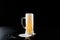 Overflowing mug of fresh cold light beer with froth flowing on the table and beautiful spilled beer on a black background with