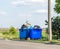 Overflowing waste cans with garbage bags, cardboard boxes and other garbage near the road. One waste can is broken