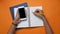 Overhead shot of woman holding smart phone and making note on empty notebook on orange background.