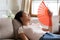 Overheated exhausted Asian woman waving paper fan, sitting on couch