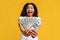 Overjoyed black woman holding money cash in hands, standing over yellow studio background, selective focus