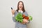 Overjoyed happy brown haired young woman embraces bouquet of fresh vegetables wearing striped casual shirt isolated over gray