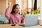 Overjoyed teen girl sitting at desk with laptop and making YES gesture, achieving success, getting good news