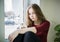 Overweight teenager girl with long blond hair is sitting by the window on the windowsill with a phone in her hand. The concept of