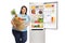 Overweight woman with a paper bag filled with fruit and vegetables leaning against an open fridge