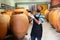 Owner in protective mask of winery standing with wine in wooden barrels cellar