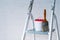 A paintbrush and bucket with a red lid stand on a metal stepladder against a white wall decorated with bricks.