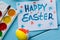 Painted Easter egg, paints, paintbrush and note with title written by paint `Happy Easter` on the blue colored wooden background