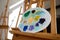 Paints colors mix on plastic palette lying on wooden easel, selective focus