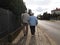 A pair of elderly people walk along the sidewalk along the road holding hands. Grandfather and grandmother on a walk in a