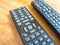 A pair of TV remotes laying next to each other. Two Television Remote Controls Side by Side on a Wooden Table. Partial