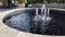 Panning footage of rippling water in a round water fountain in the garden with five water spouts spraying water and people walking