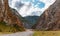 Panorama of road to the gorge of mountains on background of beautiful snowy mountain peaks in cloudy weather. Mountain road in the